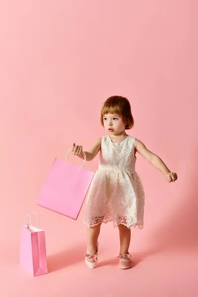 Petite fille en robe blanche tenant des sacs à provisions roses sur un fond rose. — Photo de stock