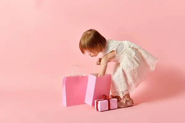 Adorable girl in white dress holding pink shopping bags against a pink background. — Stock Photo
