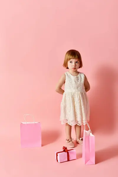 Adorable girl in white dress with gift bags against pink backdrop. — Stock Photo