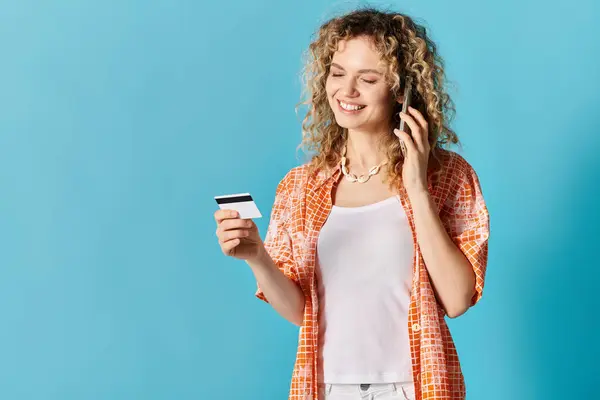 Young woman with curly hair holding credit card and talking on phone, all against vibrant backdrop. — Stock Photo