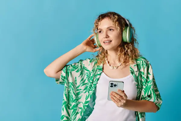 Woman with curly hair, wearing headphones, holding phone on blue background. — Stock Photo