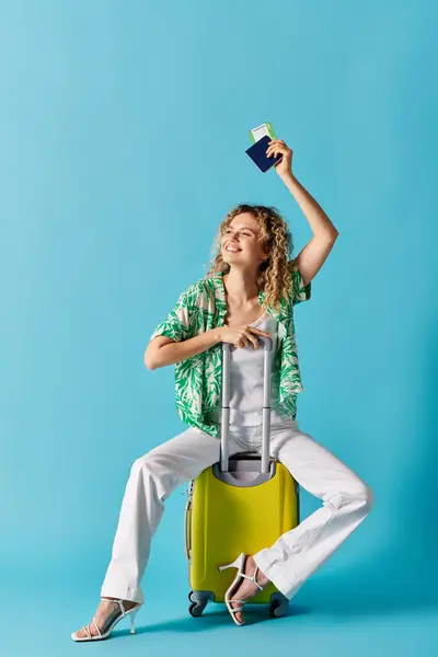 Mujer elegante con el pelo rizado sentado en la maleta amarilla vibrante, la celebración de pasaporte. - foto de stock