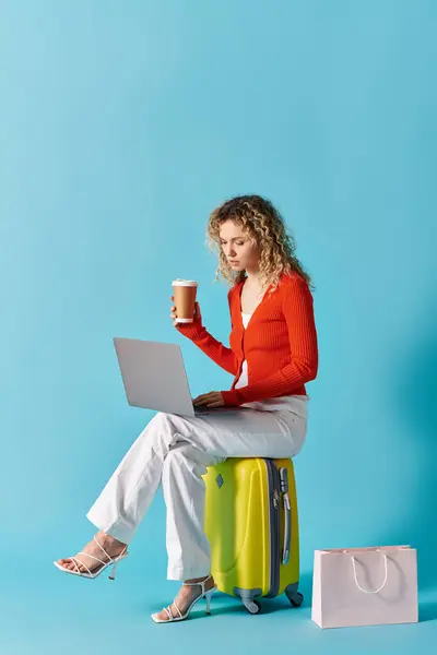 A woman with curly hair sits on a suitcase, sipping coffee and working on a laptop. — Stock Photo