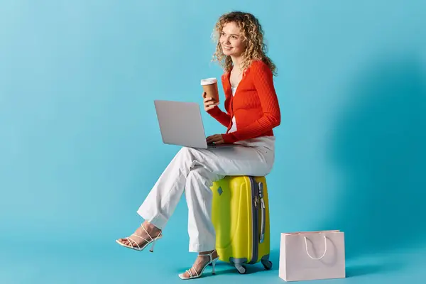 Curly-haired woman sitting on suitcase, typing on laptop, and enjoying coffee. — Stock Photo