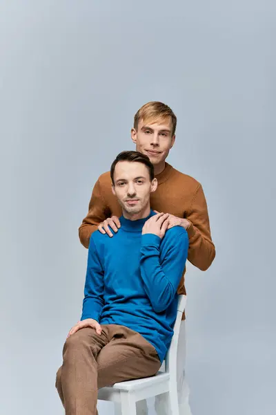 Two loving gay men in casual attire pose for a picture on a white chair against a gray backdrop. — Stock Photo