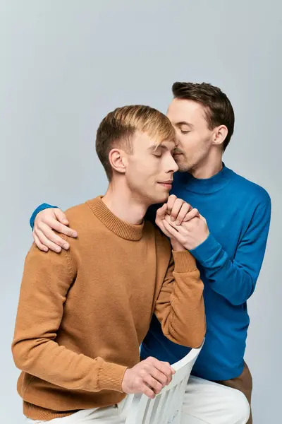 Two men, a loving gay couple, sitting together on a chair in casual attires against a gray backdrop. — Stock Photo