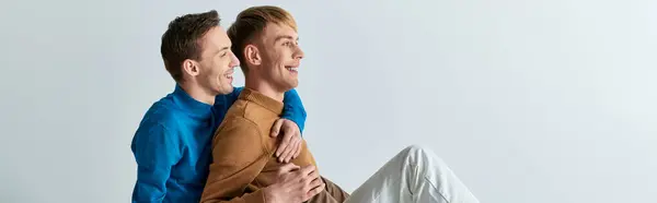 Two men in casual attire sitting lovingly intertwined on a gray backdrop. — Stock Photo