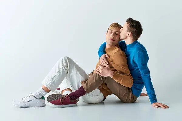 Ein liebevolles schwules Paar sitzt in lässiger Kleidung vor grauem Hintergrund auf dem Boden. — Stockfoto