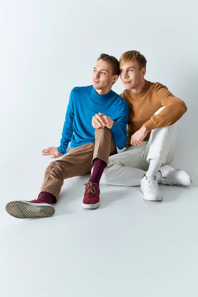 A loving gay couple in casual attires sit closely on a gray backdrop. — Stock Photo