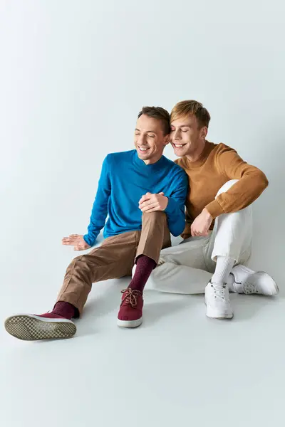 A loving gay couple in casual attires sitting closely together on a gray backdrop. — Stock Photo