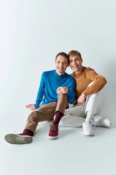 A loving gay couple in casual attire sitting closely next to each other on a gray backdrop. — Stock Photo