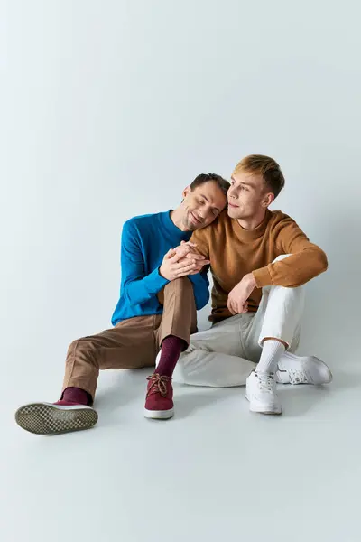 Two men, a loving gay couple, sitting next to each other in casual attire on a gray backdrop. — Stock Photo