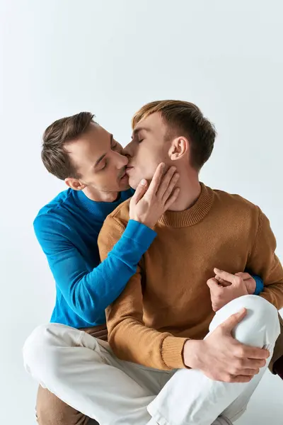 Two men with casual attire sitting close together, showcasing love and companionship against a gray backdrop. — Stock Photo