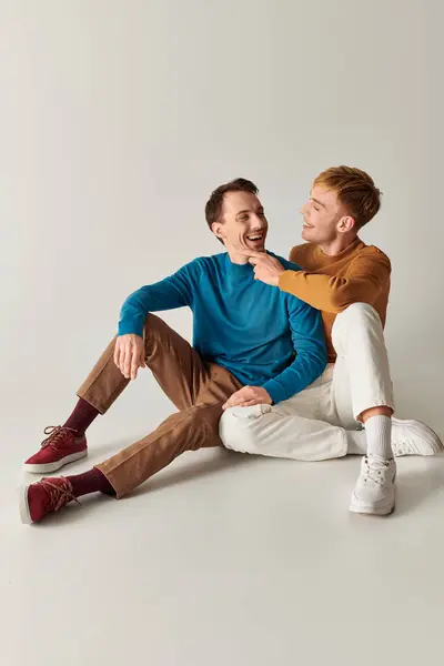 Two men sitting on the ground, laughing happily in casual attire against a gray backdrop. — Stock Photo