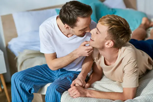 A loving gay couple in casual attire sits together on a bed. — Stock Photo