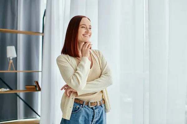 A young woman with red hair standing gracefully in front of a window in a modern apartment. — Stock Photo