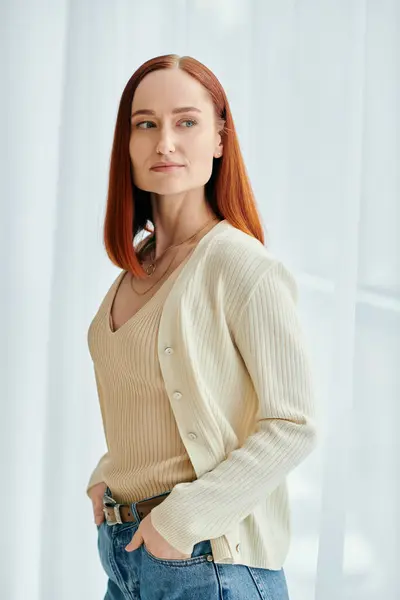 A redheaded woman in jeans and a cardigan strikes a pose in front of a window with natural light shining in. — Stock Photo