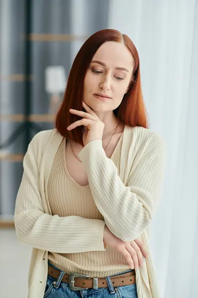 Une femme rousse se tient gracieusement devant une fenêtre, la lumière du soleil illuminant ses serrures ardentes. — Photo de stock