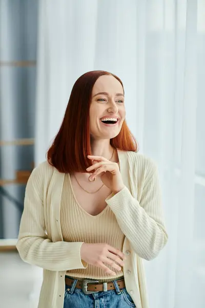 A red-haired woman laughs joyfully in front of a window with the setting sun casting a warm glow. — Stock Photo