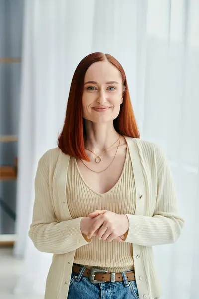 A red-haired woman standing gracefully in front of a window, basking in natural light. — Stock Photo