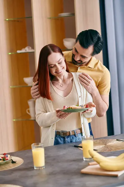 A beautiful adult couple, a redhead woman, and a bearded man, enjoying breakfast in a modern kitchen. — Stock Photo