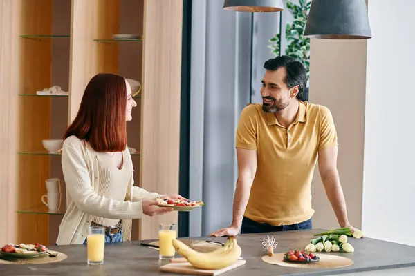 Una donna rossa e un uomo barbuto in piedi al bancone della cucina moderna, godendo di tempo di qualità insieme. — Foto stock
