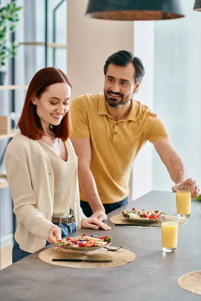 Una hermosa pareja de adultos, una mujer pelirroja y un hombre barbudo, preparando comida en una cocina moderna, disfrutando de un tiempo de calidad juntos en casa. - foto de stock