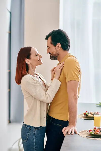 Un uomo barbuto e una donna rossa si abbracciano in una cucina calda, godendo di un momento di intimità nel loro appartamento moderno. — Foto stock