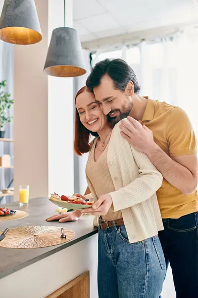 A redhead woman and bearded man in a modern kitchen, holding a plate of delicious food, enjoying quality time together. — Stock Photo
