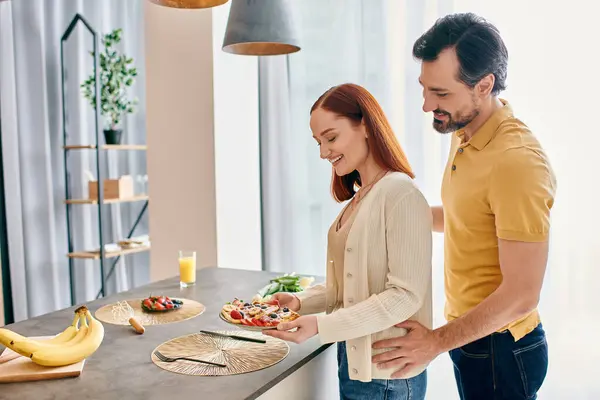 Uma mulher ruiva e um homem barbudo estão preparando alegremente comida juntos em uma cozinha moderna, unindo-se sobre a arte de cozinhar.. — Fotografia de Stock