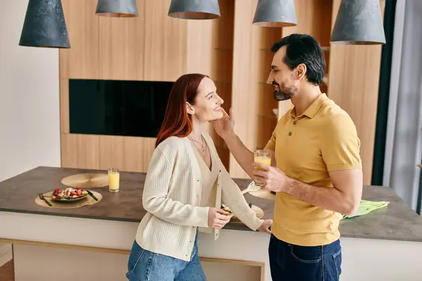 A beautiful adult couple, a redhead woman and bearded man, stand together in a modern kitchen, embracing and enjoying quality time. — Stock Photo