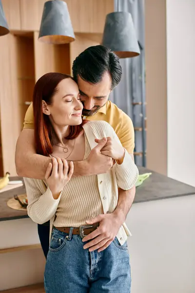 Рыжая женщина и бородатый мужчина нежно обнимаются на своей современной кухне, разделяя момент любви и связи. — стоковое фото