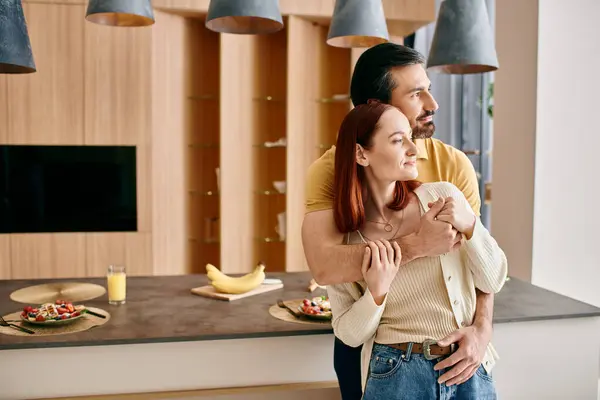 Redheaded woman and bearded man in a warm embrace in a modern kitchen, showing affection and connection. — Stock Photo
