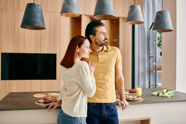 Una mujer pelirroja y un hombre barbudo se unen en una cocina elegante, disfrutando de un momento de unión en su apartamento moderno. - foto de stock