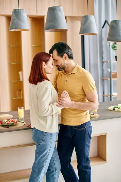A bearded man and a redhead woman are happily dancing together in a sunlit modern kitchen. — Stock Photo