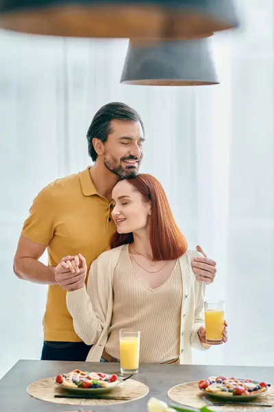 Young redhead woman and bearded man standing affectionately by kitchen table in modern apartment. — Stock Photo