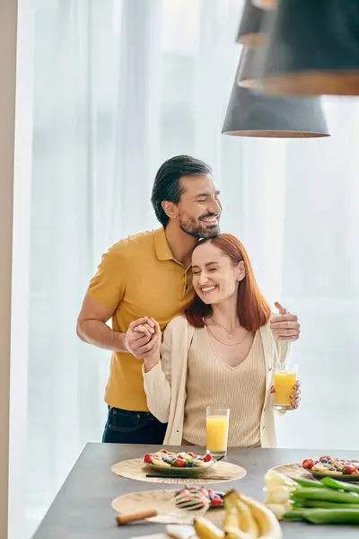 Una donna rossa e un uomo barbuto godono di una mattinata rilassante nella loro cucina moderna con bicchieri di succo d'arancia fresco. — Foto stock