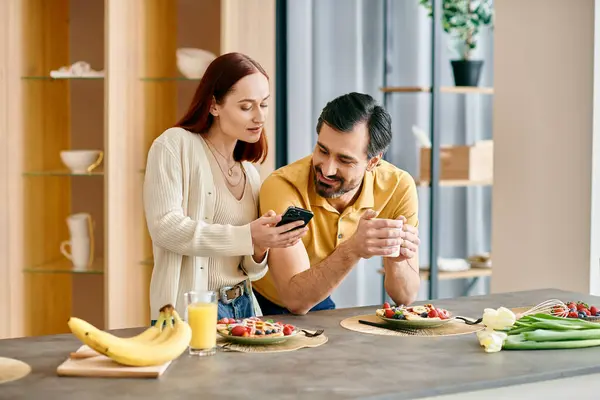 Una hermosa pareja de adultos, una mujer pelirroja y un hombre barbudo, disfrutan de una comida juntos mientras están comprometidos con sus teléfonos. - foto de stock