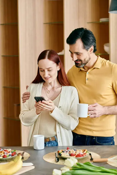 Руда жінка і бородатий чоловік зосереджені на екранах свого телефону, стоячи на сучасній кухні. — стокове фото