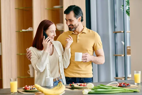 A redhead woman and a bearded man talk on the phone while enjoying breakfast in a modern kitchen. — Stock Photo