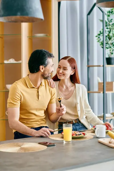 Uma mulher ruiva e um homem barbudo sentados em uma mesa em uma cozinha moderna, compartilhando um momento tranquilo juntos. — Fotografia de Stock