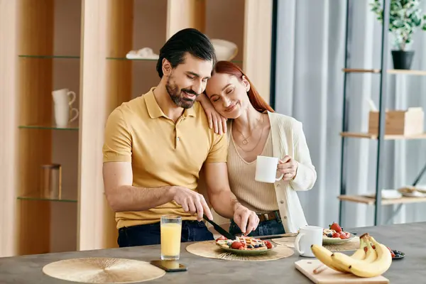 A redhead woman and a bearded man are sitting at a kitchen table, eating breakfast together in a modern apartment. — Stock Photo