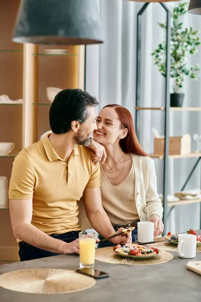 Una hermosa pareja adulta, una pelirroja y un hombre barbudo, sentados a la mesa, desayunando en un apartamento moderno. - foto de stock