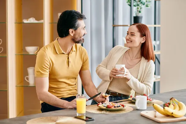 A bearded man and a redhead woman enjoying breakfast together at their kitchen table in a modern apartment. — Stock Photo