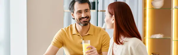 Uma mulher ruiva e um homem barbudo sentados em uma mesa, desfrutando de um refrescante copo de suco de laranja em seu apartamento moderno. — Fotografia de Stock