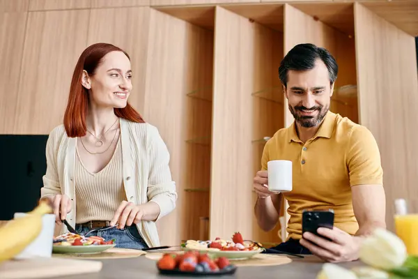 A redheaded woman and a bearded man sit at a table, happily eating breakfast in their modern apartment. — Stock Photo