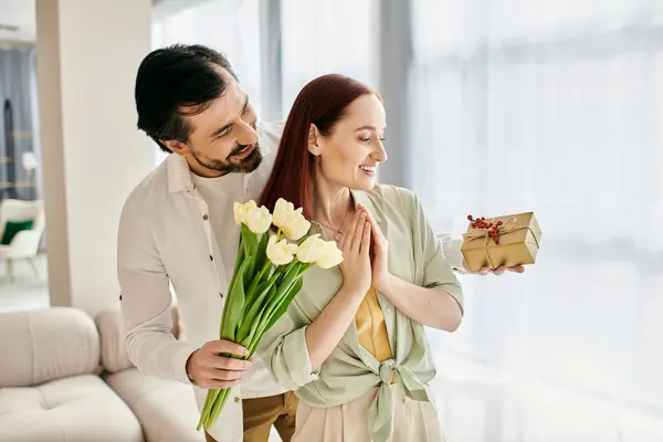 Um homem barbudo apresenta ternamente um buquê de tulipas a uma mulher ruiva em um ambiente de apartamento moderno, incorporando amor e união. — Fotografia de Stock