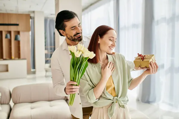 A beautiful adult couple, a redhead woman, and a bearded man, standing in a modern living room holding a gift box. — Stock Photo