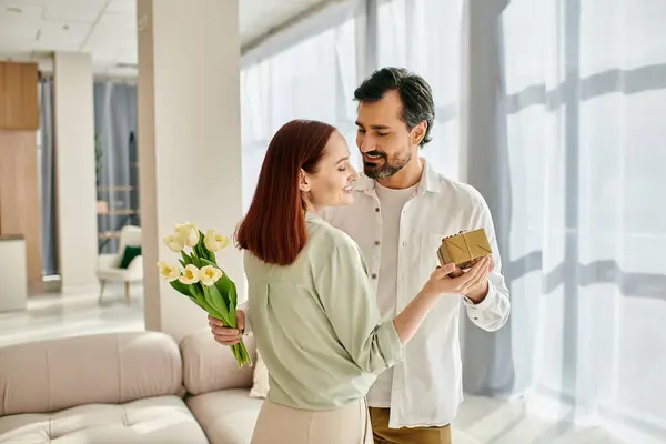 A bearded man presents a gift to a redhead woman in a modern living room, creating a heartfelt moment of connection and love. — Stock Photo