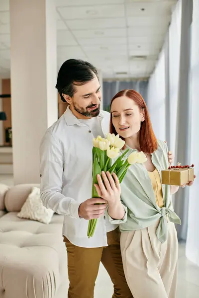 A redhead woman and bearded man hold flowers in a modern living room, enjoying quality time together. — Stock Photo